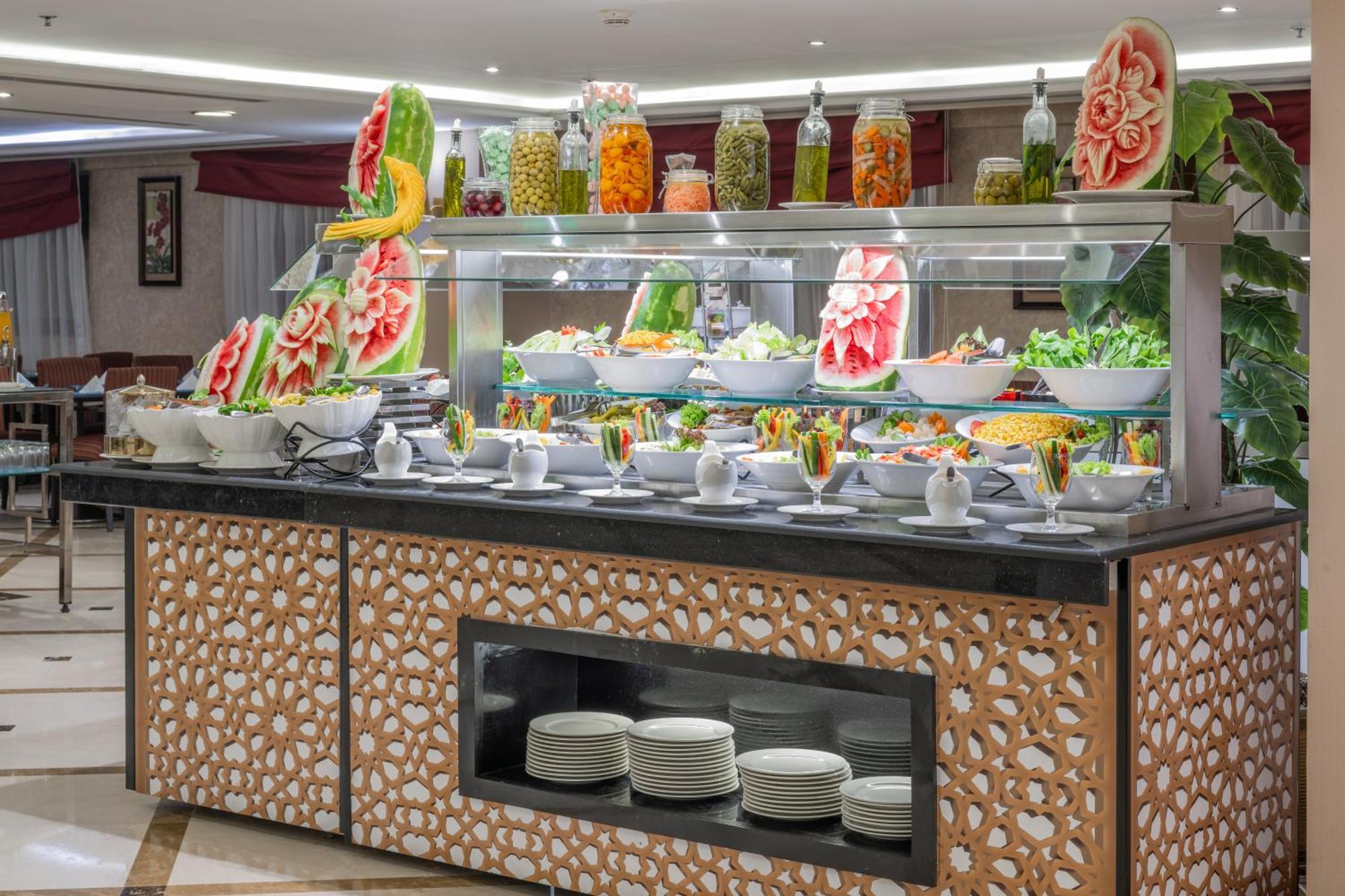 Al Aqeeq Madinah Hotel Medina Exterior photo The photo shows a buffet display that features an array of colorful food items. In the foreground, there are several large bowls filled with various salads and dishes. The presentation is visually appealing, with decorative watermelon carvings and an