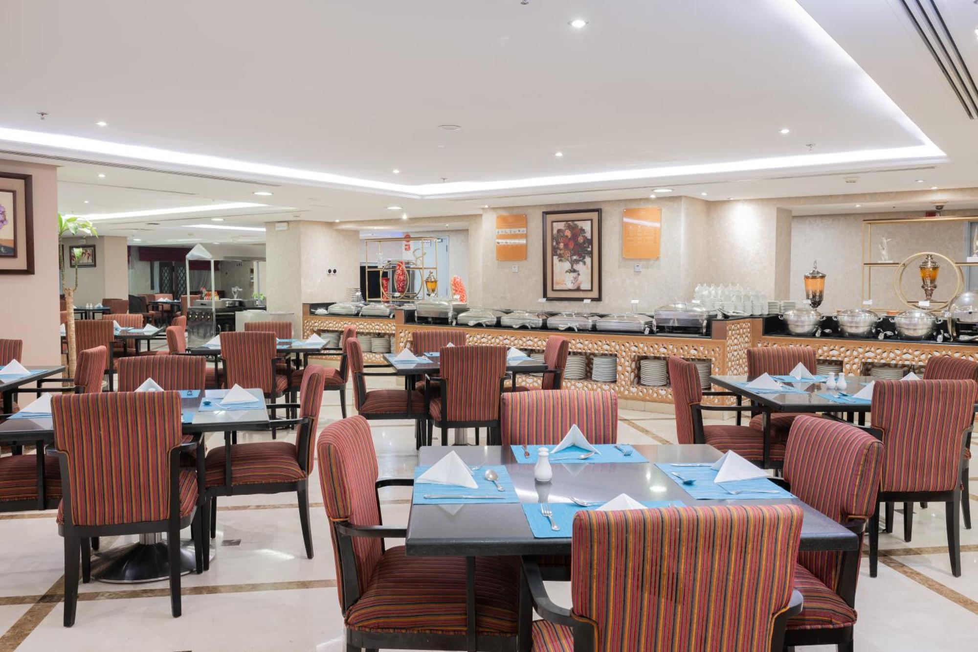 Al Aqeeq Madinah Hotel Medina Exterior photo The photo shows the interior of a restaurant. It features multiple tables arranged neatly, each with patterned chairs and set with blue and white table settings, including folded napkins. The lighting is bright and modern, creating a welcoming atmosp