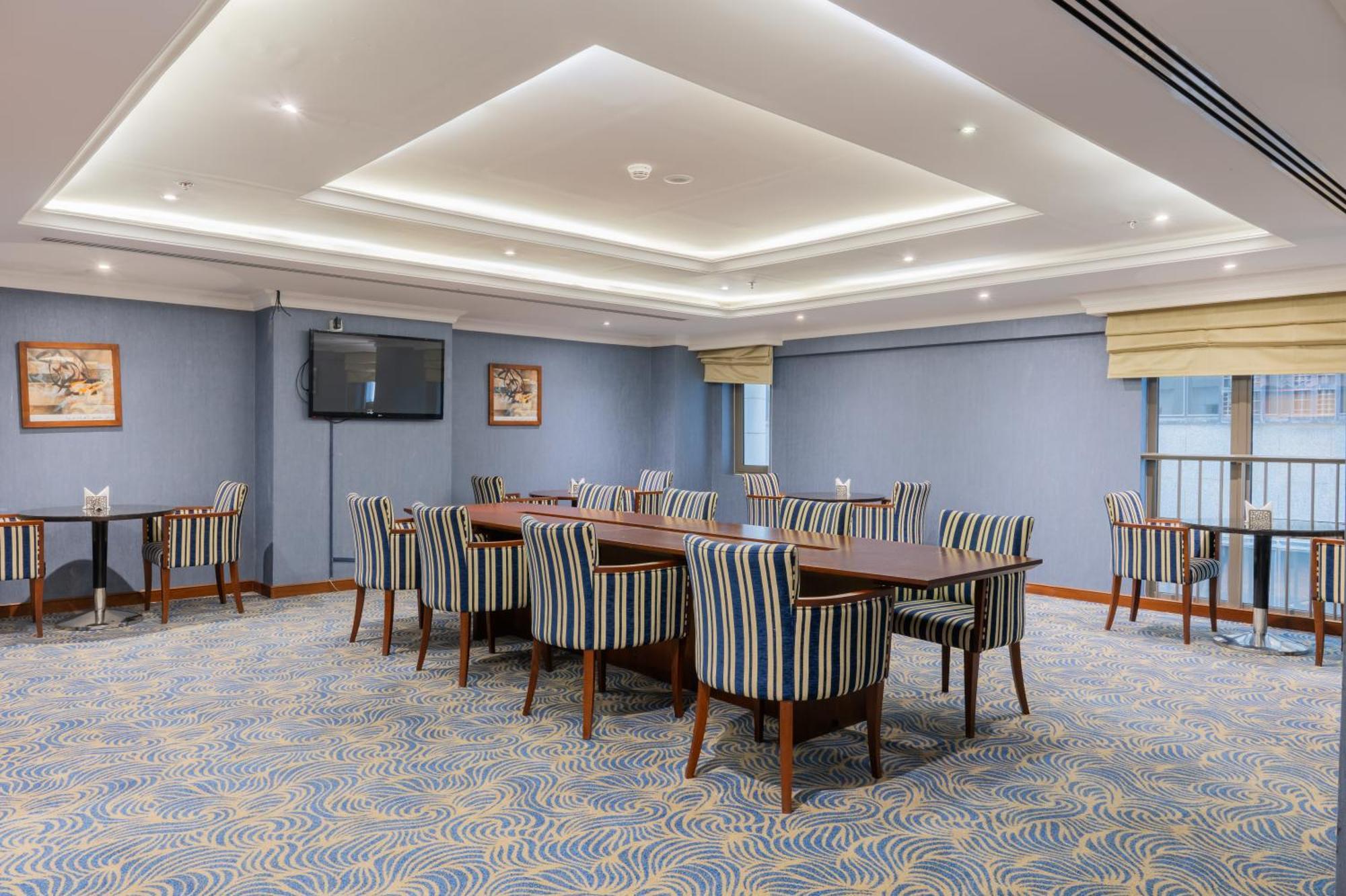 Al Aqeeq Madinah Hotel Medina Exterior photo The photo shows a spacious conference or meeting room. It features a light blue color scheme with a patterned carpet. There are several rectangular tables arranged in a group, surrounded by striped chairs. The room is well-lit with modern ceiling lig
