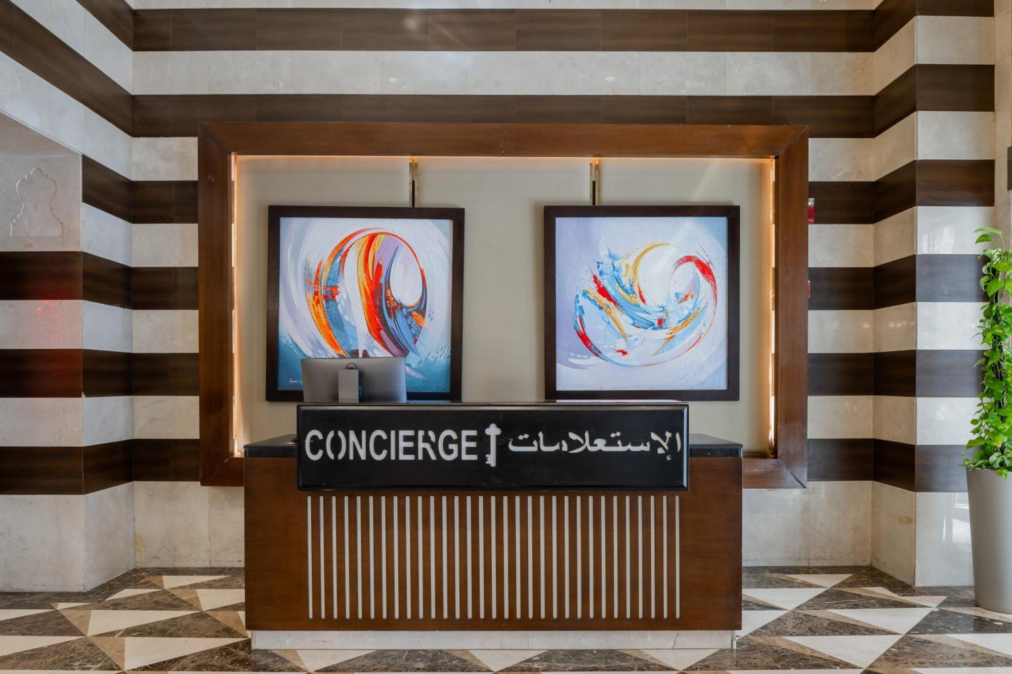 Al Aqeeq Madinah Hotel Medina Exterior photo The image shows a concierge desk in a hotel lobby. The desk is made of wood with a striped design and features two large, colorful abstract paintings on the wall behind it. The sign on the desk reads "CONCIERGE" in English and has Arabic text below i