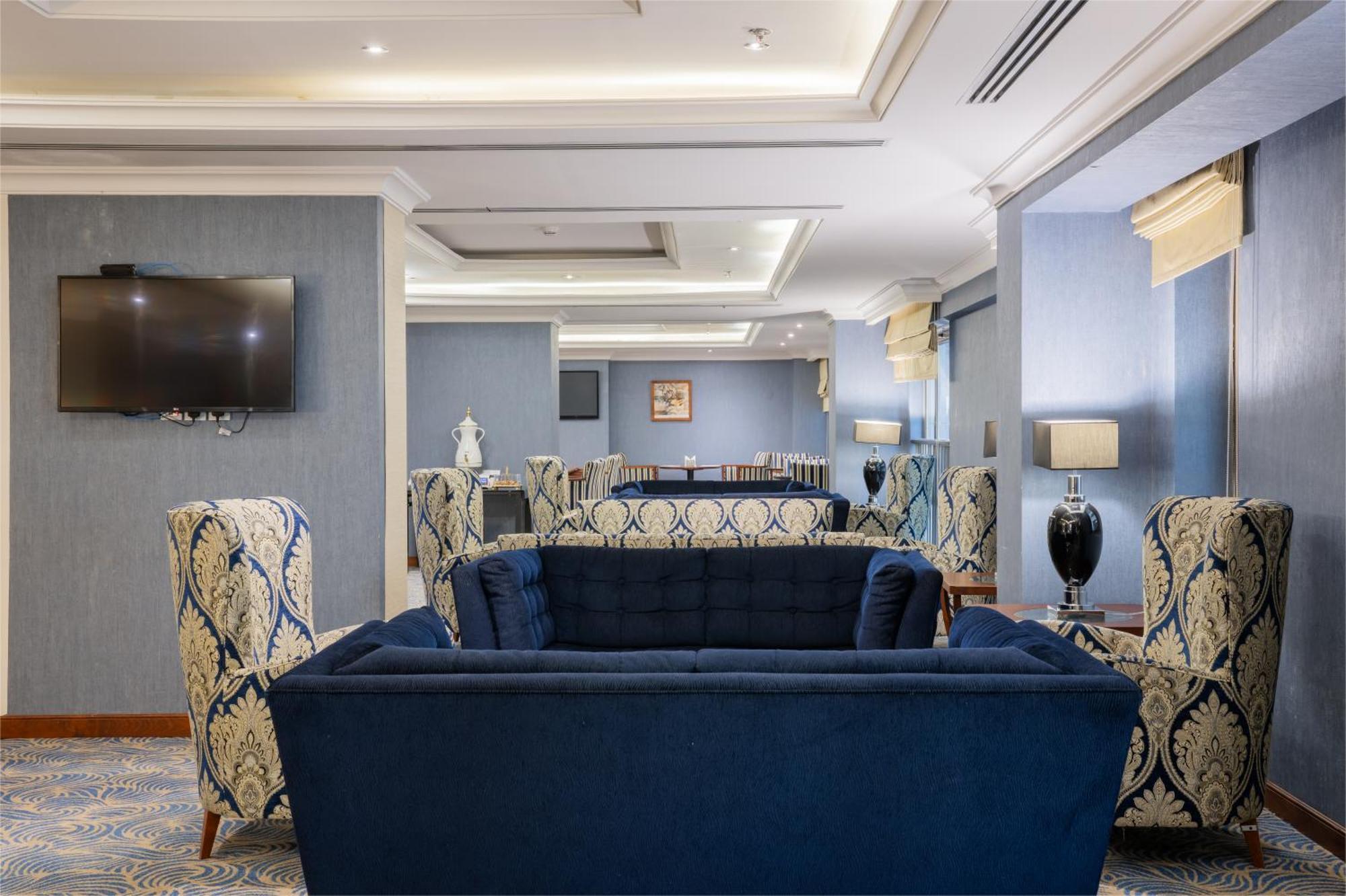 Al Aqeeq Madinah Hotel Medina Exterior photo The photo shows a stylish and modern lounge area. There are two patterned armchairs with blue and cream designs, positioned next to a deep blue couch. The room has a light blue wall with a flat-screen TV mounted on one side. Soft lighting from stylis