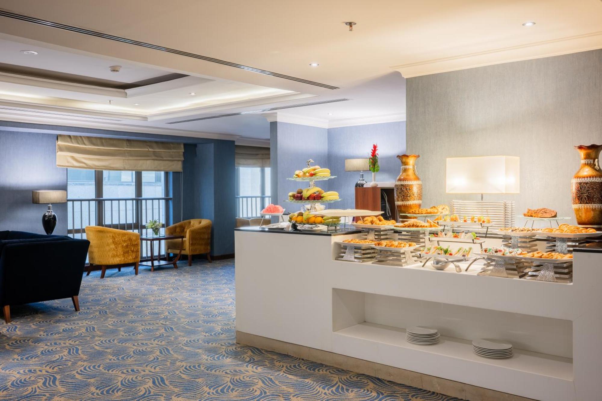 Al Aqeeq Madinah Hotel Medina Exterior photo The photo shows a modern dining area, likely in a hotel or restaurant. On the right side, there is a buffet table with various food items displayed, including pastries and desserts. Behind the table, a decorative fountain is visible. The room feature