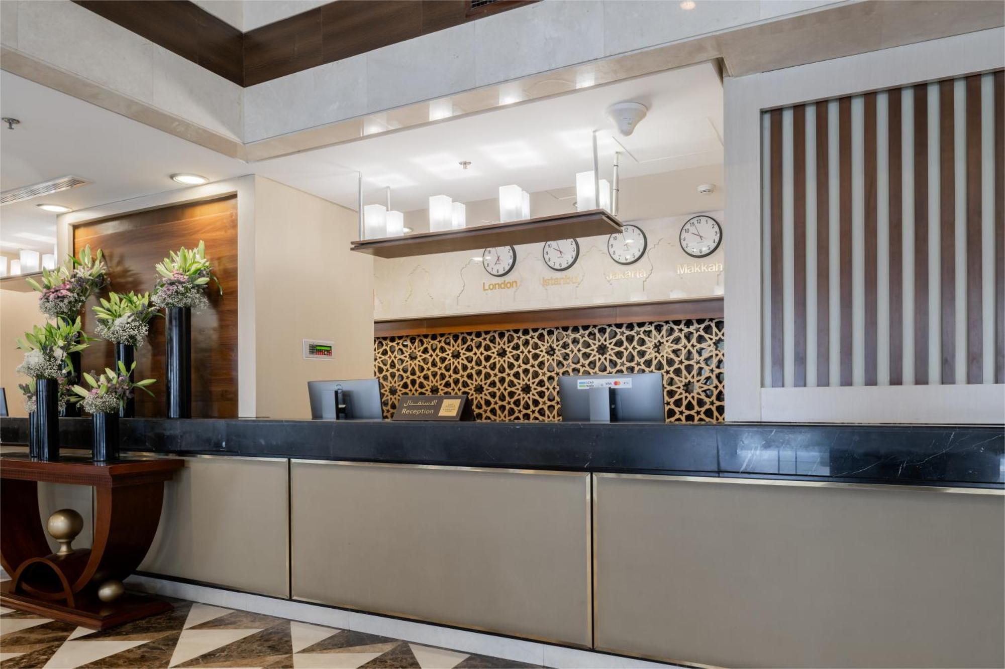 Al Aqeeq Madinah Hotel Medina Exterior photo The photo shows the interior of a hotel reception area. The reception desk is designed with a sleek, modern look featuring a dark countertop and a light-colored front. Above the desk, there are several clocks displaying different times, suggesting an