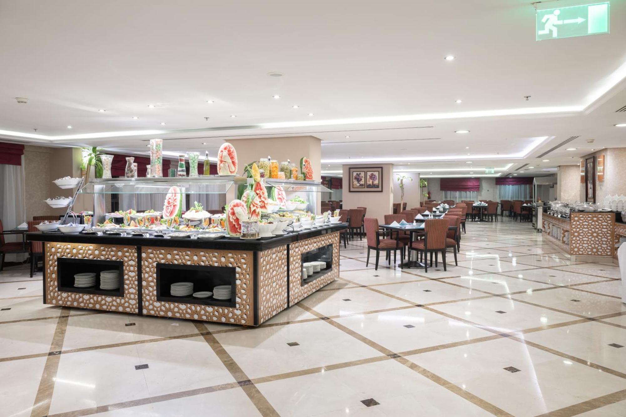 Al Aqeeq Madinah Hotel Medina Exterior photo The photo shows a spacious restaurant interior with a modern design. In the foreground, there is a large buffet counter filled with various food items, beautifully arranged and decorated. The counter features a backlit display, showcasing fruits and 