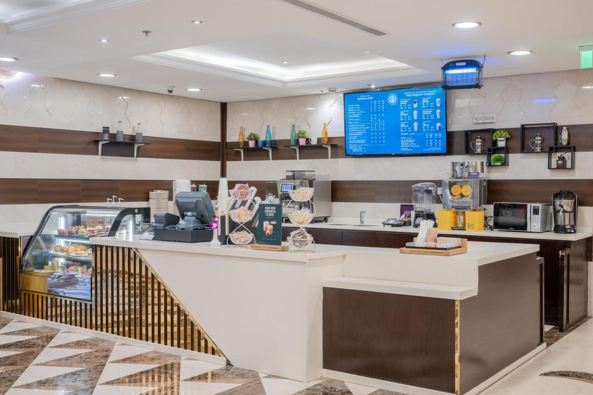 Al Aqeeq Madinah Hotel Medina Exterior photo The photo shows a modern café or coffee shop interior. There's a counter area made of smooth white and dark brown materials, featuring a cash register and display items. Behind the counter, shelves are stocked with various beverage items and equipmen