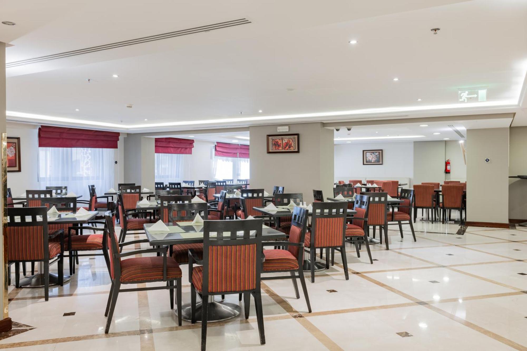 Al Aqeeq Madinah Hotel Medina Exterior photo The photo shows the interior of a restaurant or dining area. The space features several tables arranged with chairs, most of which have striped orange and black upholstery. The flooring is likely marble or tile, and the lighting is bright, creating a