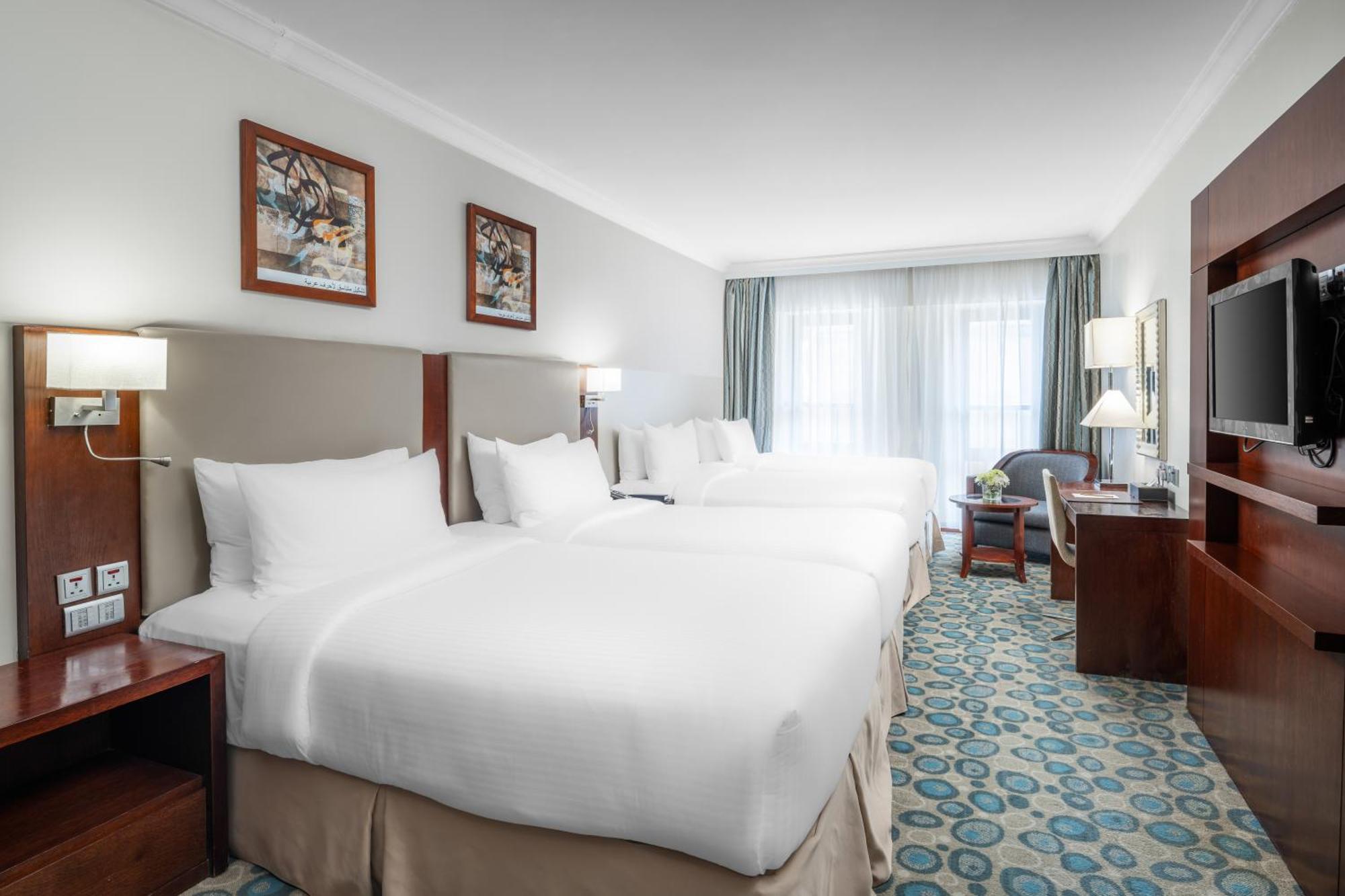 Al Aqeeq Madinah Hotel Medina Exterior photo The photo shows a hotel room featuring a modern, clean design. There are two large beds with white linens positioned side by side. Above the beds are framed artworks, and soft lighting is provided by wall sconces. To the right, there is a wooden ente