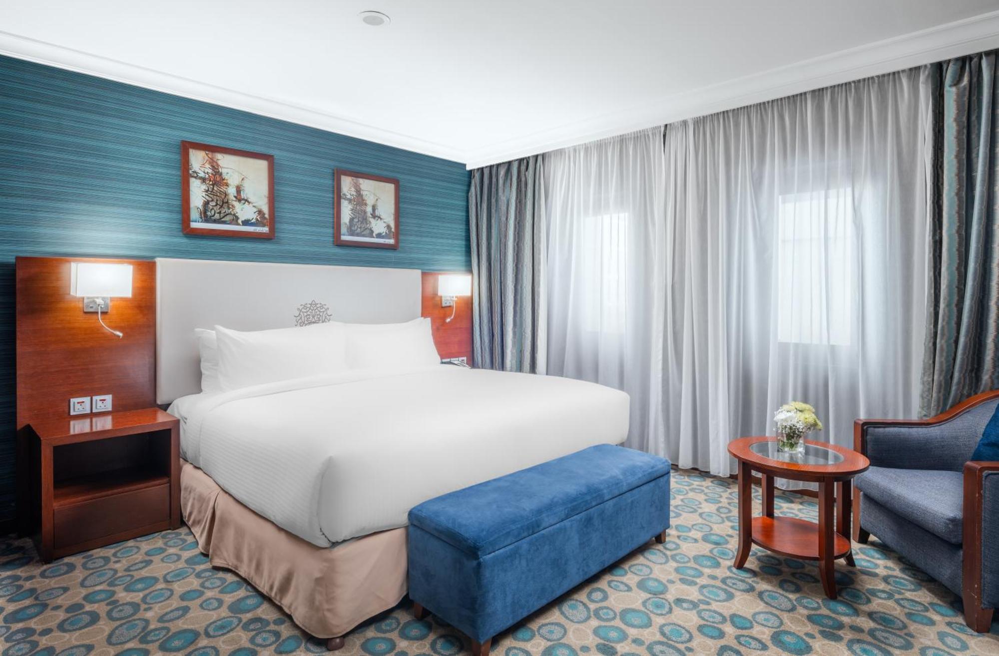 Al Aqeeq Madinah Hotel Medina Exterior photo The photo shows a hotel room featuring a large, neatly made bed with a white duvet. The walls have a light blue accent with framed artwork above the bed. There’s a blue ottoman at the foot of the bed. To one side, there’s a small wooden table with a 