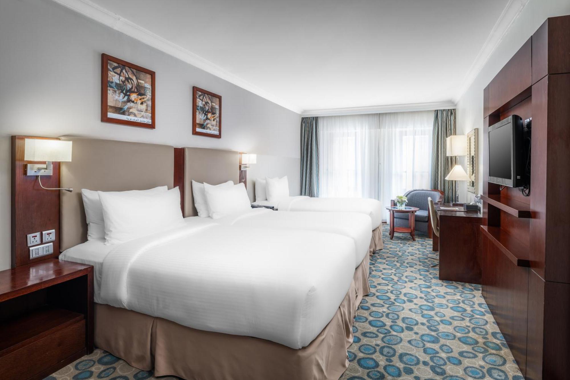 Al Aqeeq Madinah Hotel Medina Exterior photo The photo shows a well-furnished hotel room featuring two large beds with white bedding. The walls are adorned with framed artwork. There are bedside lamps next to each bed, and a flat-screen TV is mounted on the wall. A large window allows natural l