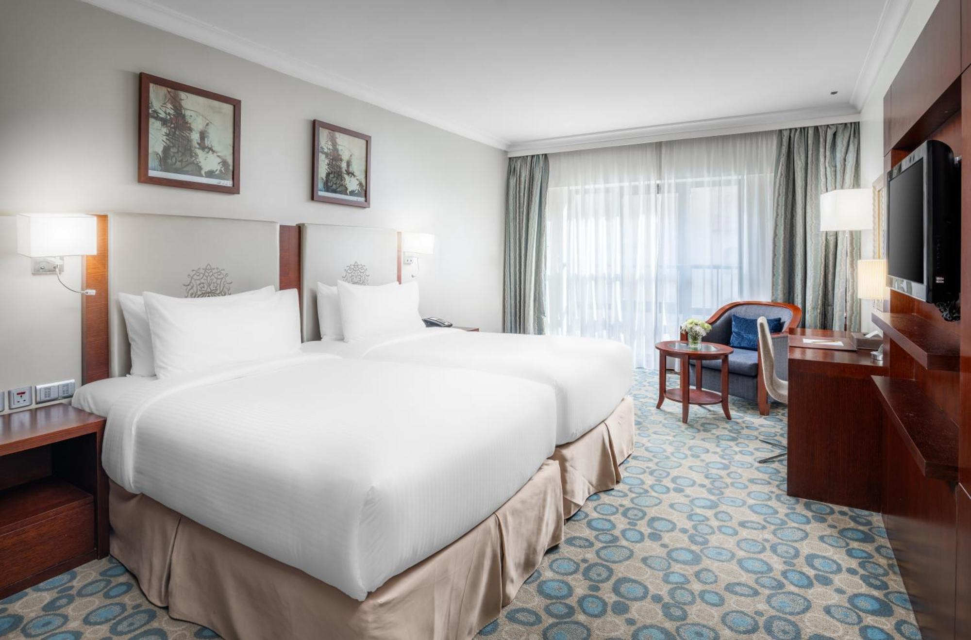Al Aqeeq Madinah Hotel Medina Exterior photo The photo shows a hotel room featuring two white beds with plush bedding. There are decorative artworks on the walls above the beds. A large window with sheer curtains allows natural light into the space, illuminating the room. The floor is covered w