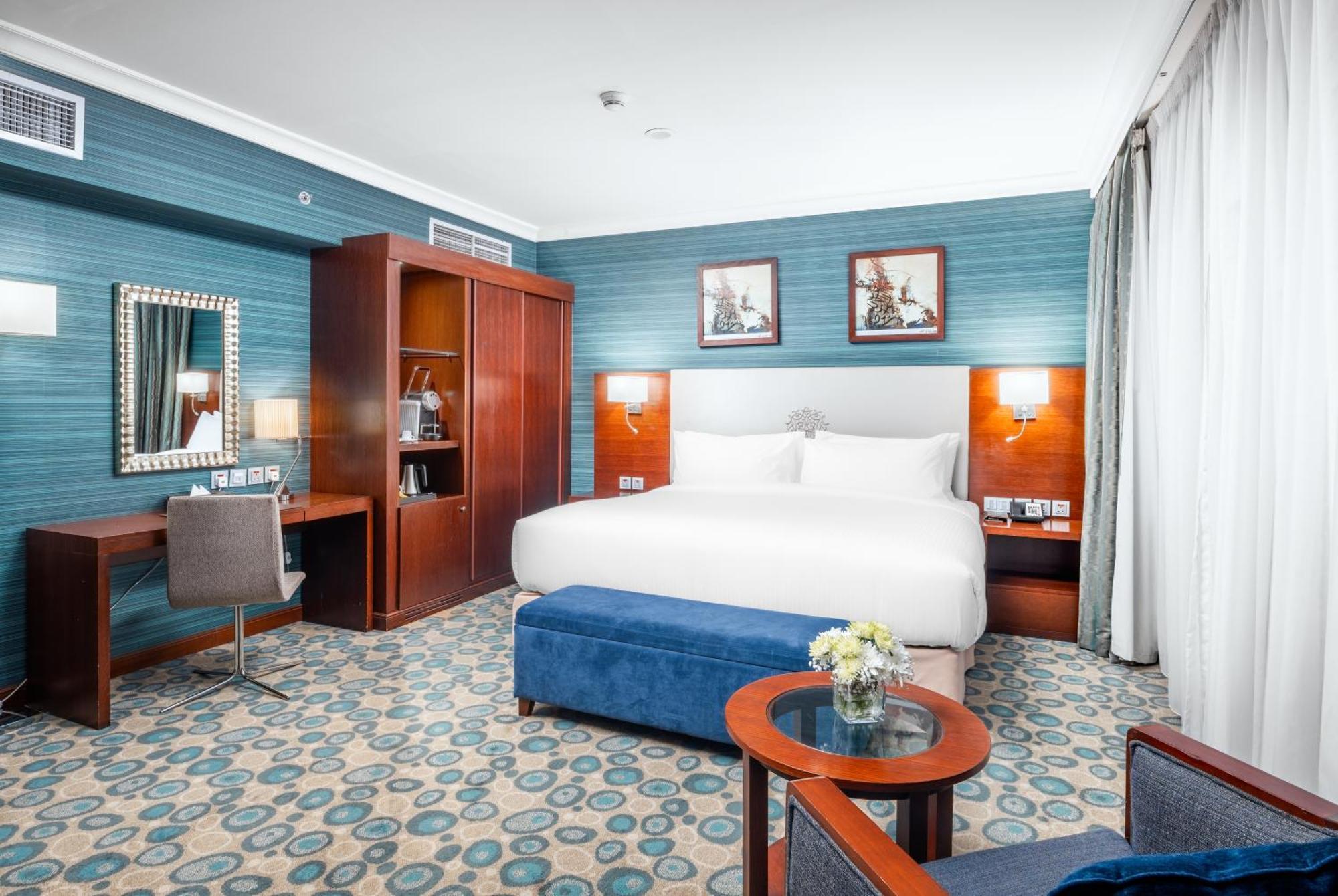 Al Aqeeq Madinah Hotel Medina Exterior photo The photo shows a well-decorated hotel room. It features a large bed with white bedding, flanked by wall-mounted lights. Behind the bed, there are two framed pictures hanging on a blue wall. To the left, there's a wooden wardrobe and a desk with a ch