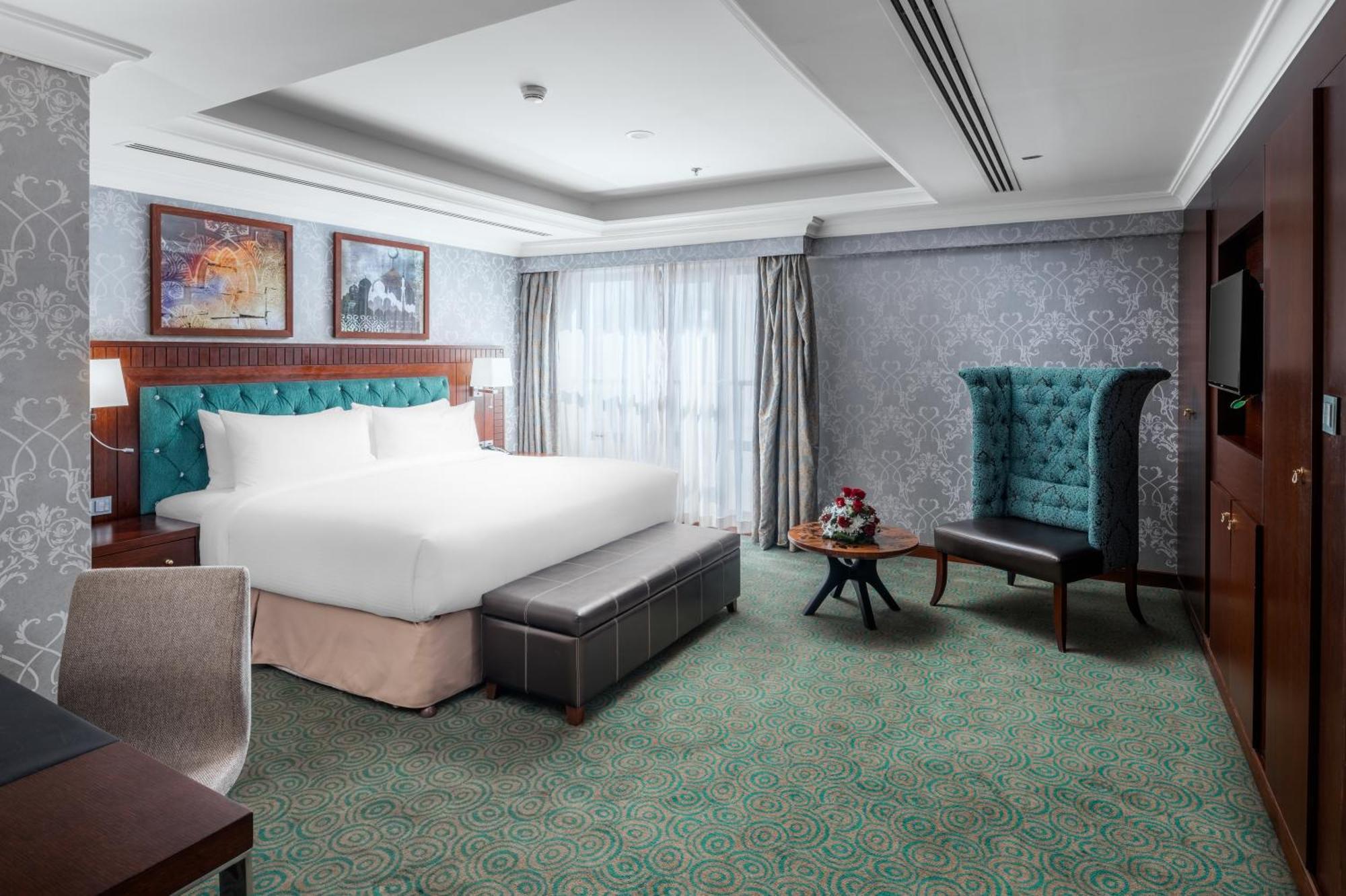 Al Aqeeq Madinah Hotel Medina Exterior photo The photo shows a well-appointed hotel room. It features a large bed with a tufted headboard in a turquoise color, complemented by decorative pillows. There is a comfortable ottoman at the foot of the bed and a small coffee table with a vase of flowe