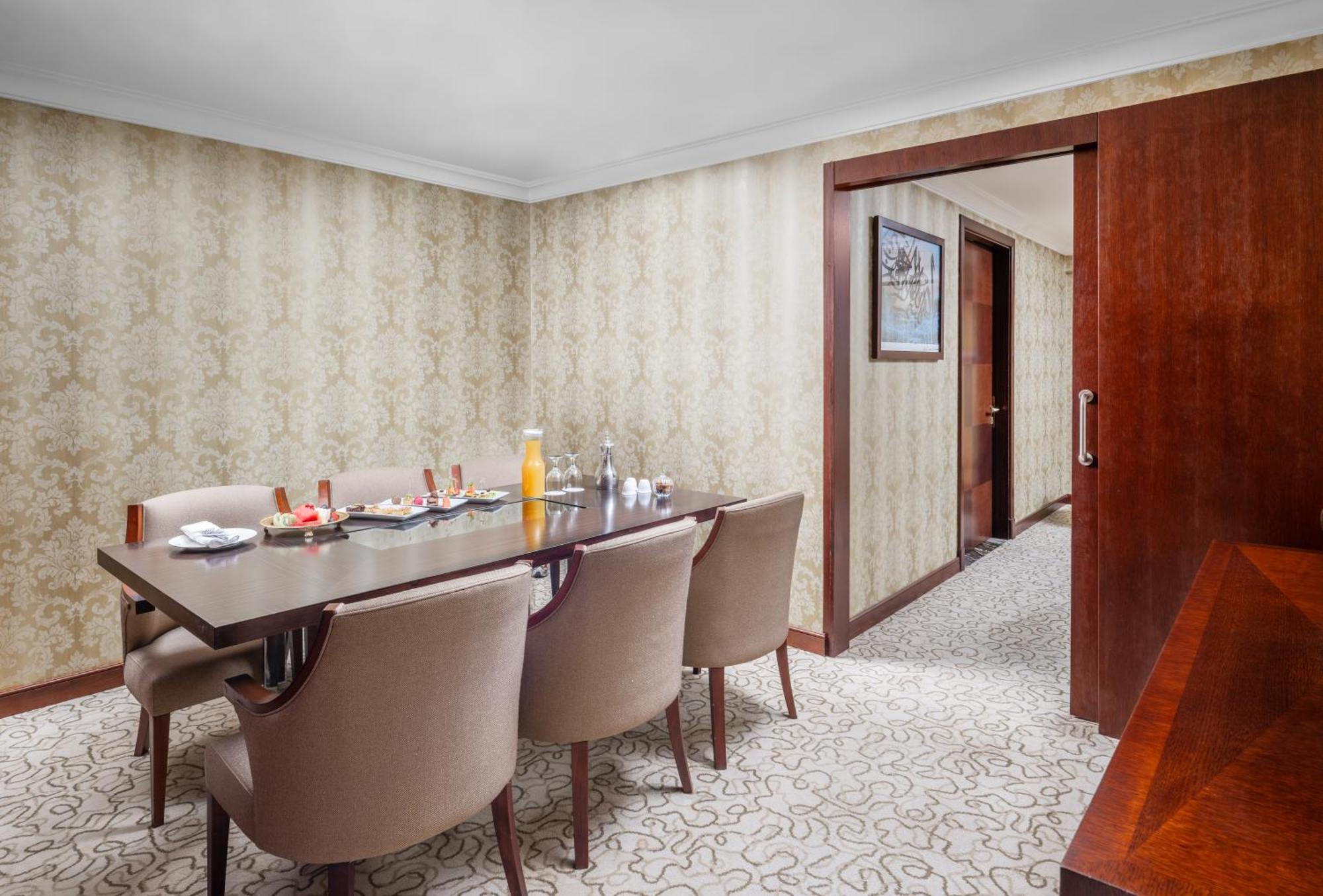 Al Aqeeq Madinah Hotel Medina Exterior photo The photo shows a dining room with a stylish and elegant design. There is a large wooden dining table surrounded by plush, beige chairs. On the table, there are various items including a bottle of orange juice, glasses, and other dining accessories. 