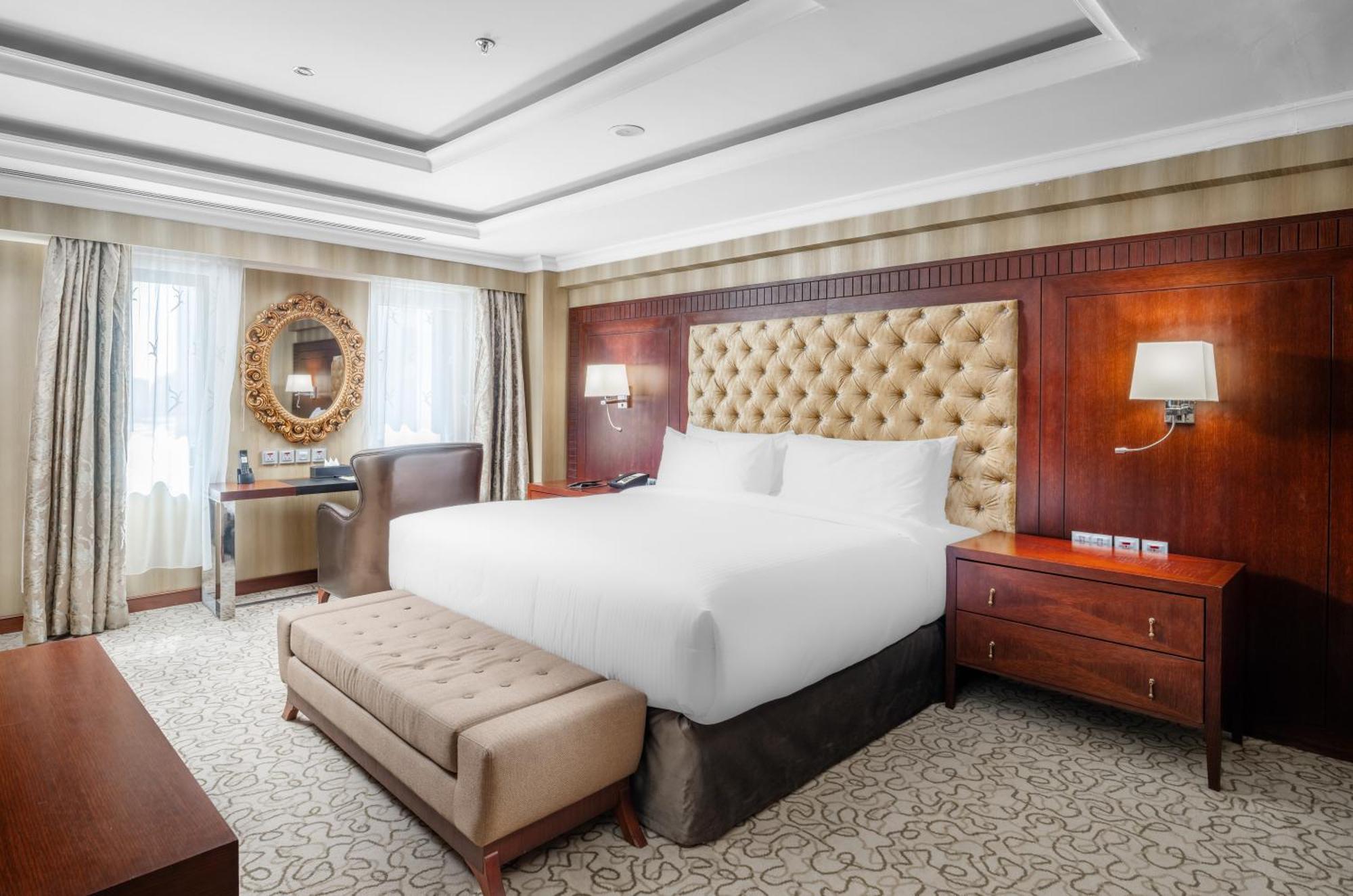 Al Aqeeq Madinah Hotel Medina Exterior photo The photo shows a well-furnished hotel room featuring a large bed with a tufted headboard. The room has a warm color palette with wooden accents and soft lighting. There are two windows with curtains that allow natural light to fill the space. Alongs