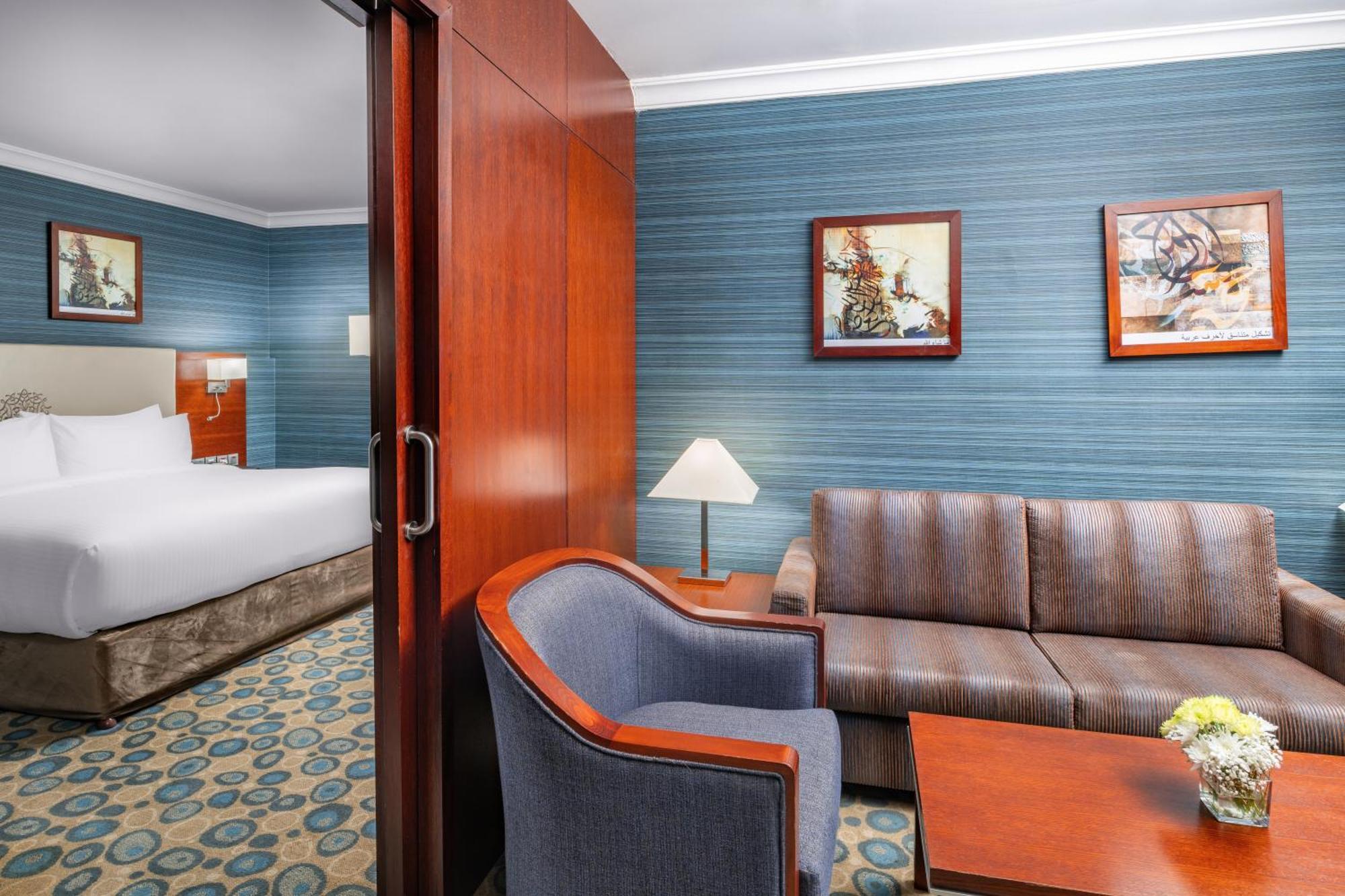 Al Aqeeq Madinah Hotel Medina Exterior photo The photo shows a well-furnished hotel room featuring a living area and a bedroom. To the left, there is a bed visible through an open doorway. The living area includes a gray upholstered chair and a brown couch, both arranged around a small table. A