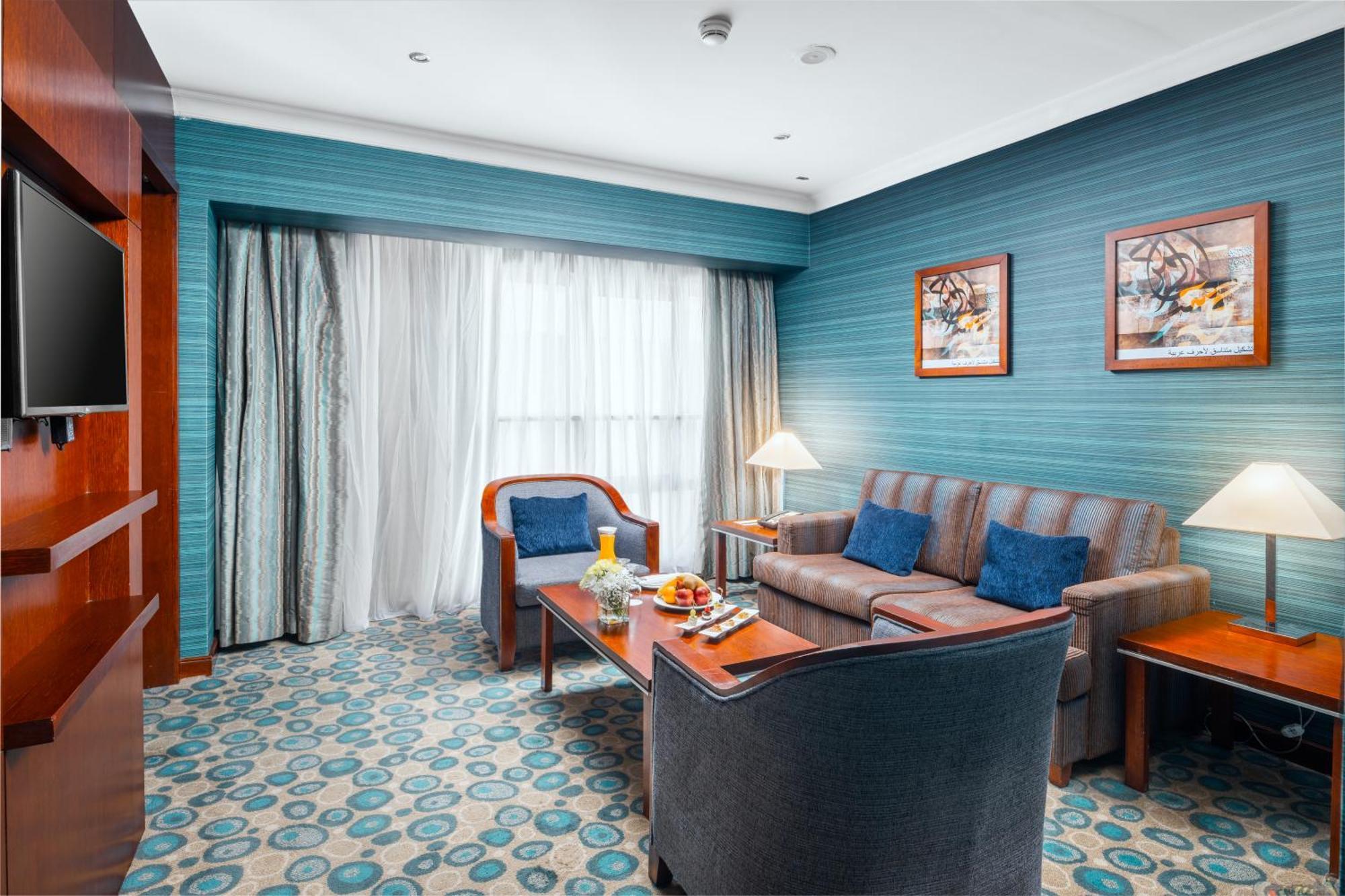 Al Aqeeq Madinah Hotel Medina Exterior photo The photo shows a cozy and modern hotel room featuring a sitting area. The walls are painted a calming teal color, complemented by a patterned carpet in shades of blue and beige. There is a comfortable sofa with cushions, two armchairs, and a wooden 