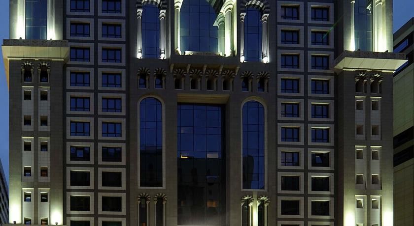 Al Aqeeq Madinah Hotel Medina Exterior photo The image shows the facade of a modern high-rise building illuminated at night. The design features large glass windows arranged in a grid pattern, along with decorative architectural elements at the top, including arches and ornate detailing. The co