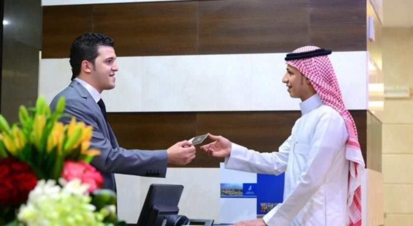 Al Aqeeq Madinah Hotel Medina Exterior photo The image depicts two individuals at a reception desk. The person on the left, dressed in a formal suit, appears to be handing a card or document to the person on the right, who is wearing traditional attire, including a white thobe and a red and whi