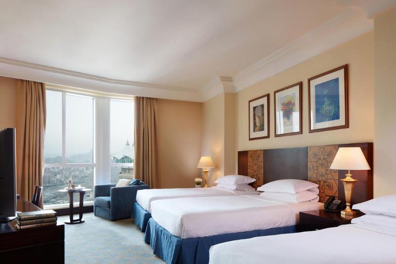 Al Aqeeq Madinah Hotel Medina Exterior photo The photo shows a hotel room featuring three beds arranged neatly. The room has a warm, inviting ambiance with beige walls and a soft carpet. Large windows allow natural light to fill the space, offering a view outside. There are curtains in a light 