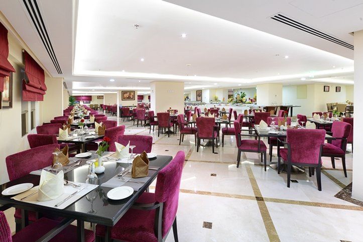 Al Aqeeq Madinah Hotel Medina Exterior photo The photo shows an elegant restaurant interior with a spacious layout. The dining area features multiple tables, each set with white tablecloths and dinnerware. The chairs are upholstered in a rich purple fabric, adding a touch of luxury to the atmos