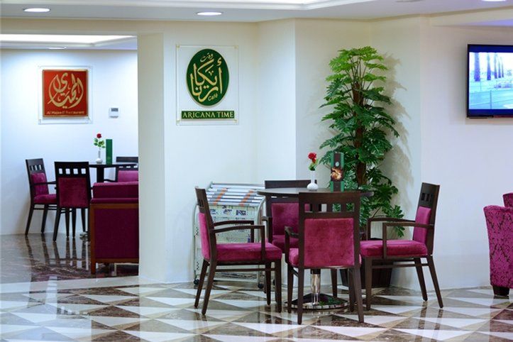 Al Aqeeq Madinah Hotel Medina Exterior photo The photo shows an interior space, likely a dining or lounge area. There are several tables with four chairs each, featuring a modern design with dark wood and plush maroon upholstery. On the walls, there are decorative signs, one of which has Arabic