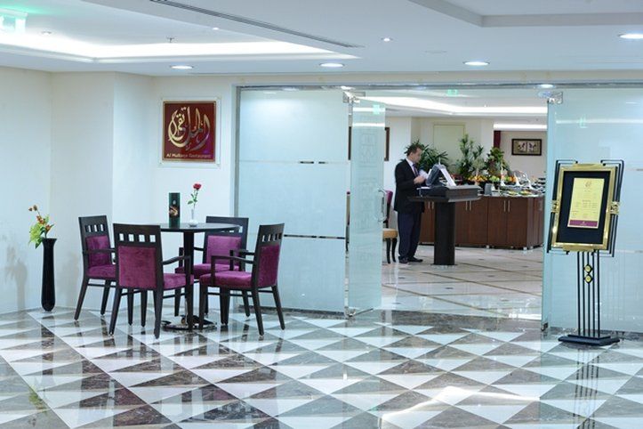Al Aqeeq Madinah Hotel Medina Exterior photo The photo shows a modern interior space, likely a lobby or reception area of a hotel or restaurant. It features a checkered floor with a combination of dark and light tiles. On the left, there is a small table set for two, adorned with a single rose 