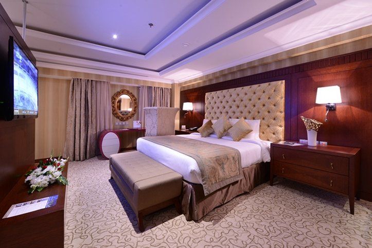 Al Aqeeq Madinah Hotel Medina Exterior photo The photo shows an elegantly designed hotel room. It features a large bed with a plush headboard and decorative pillows. On either side of the bed are bedside tables with lamps. There's a comfortable bench at the foot of the bed and a TV mounted on t